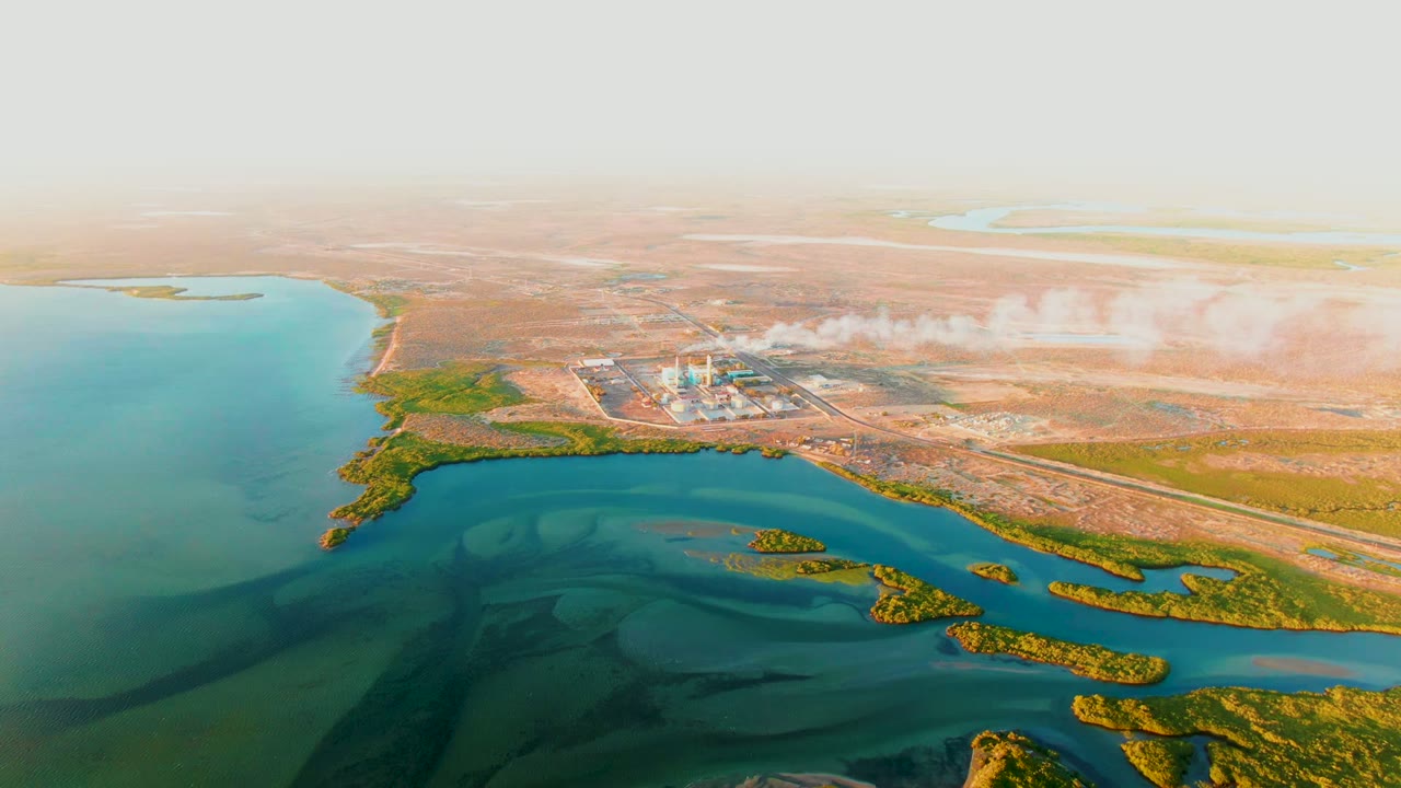 Aerial view in San Carlos, Baja California Sur showing blue-green water, low shoreline vegetation, an industrial plant, and a white smoke plume over dry land with no visible people.