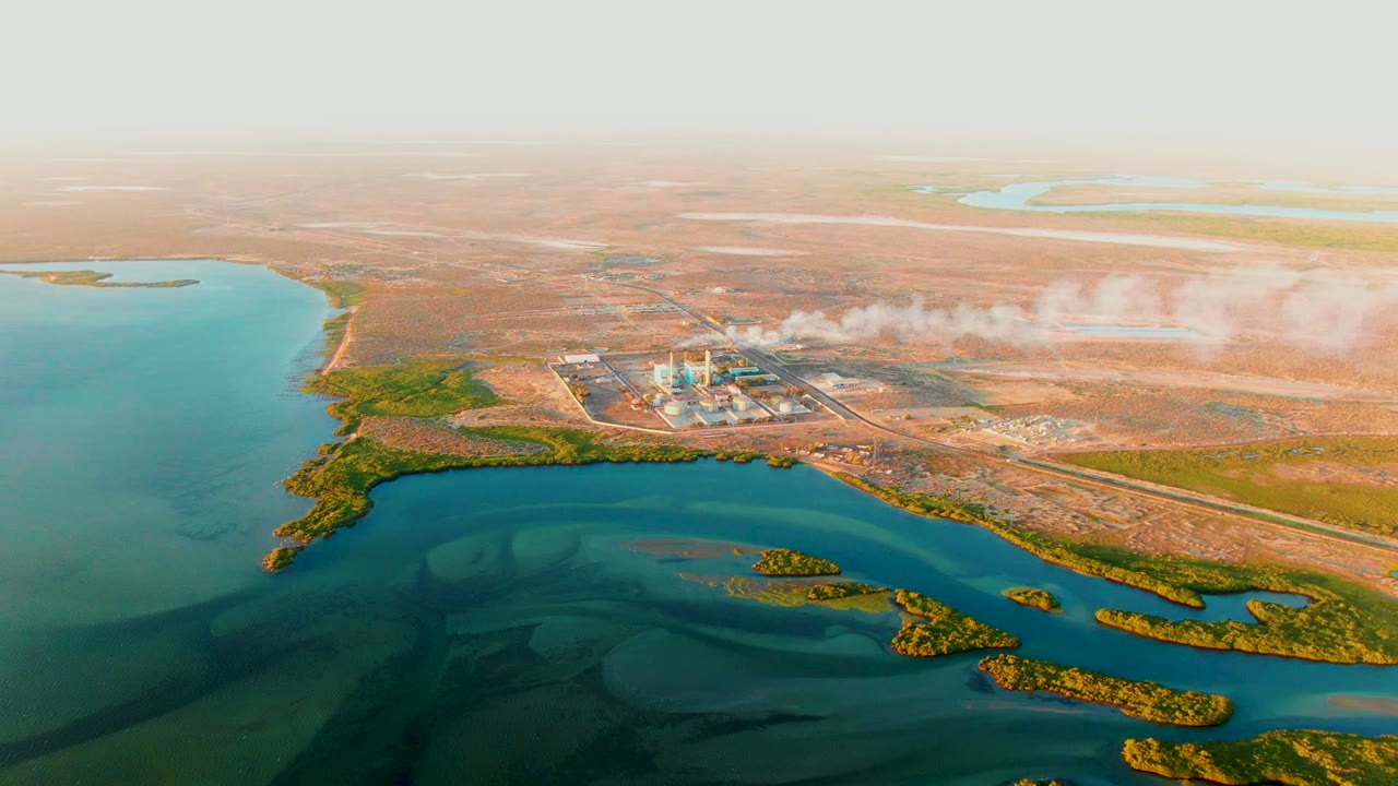 Aerial view in San Carlos, Baja California Sur where shallow blue water and small vegetated islands lead toward an industrial facility emitting a white plume, with no visible people.