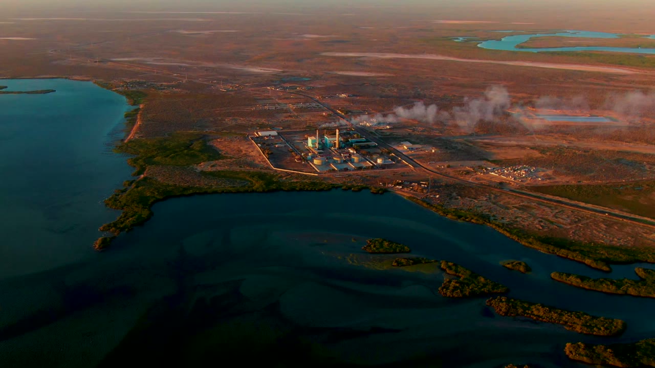 Power plant smokestacks release pale smoke over water and dry terrain.
