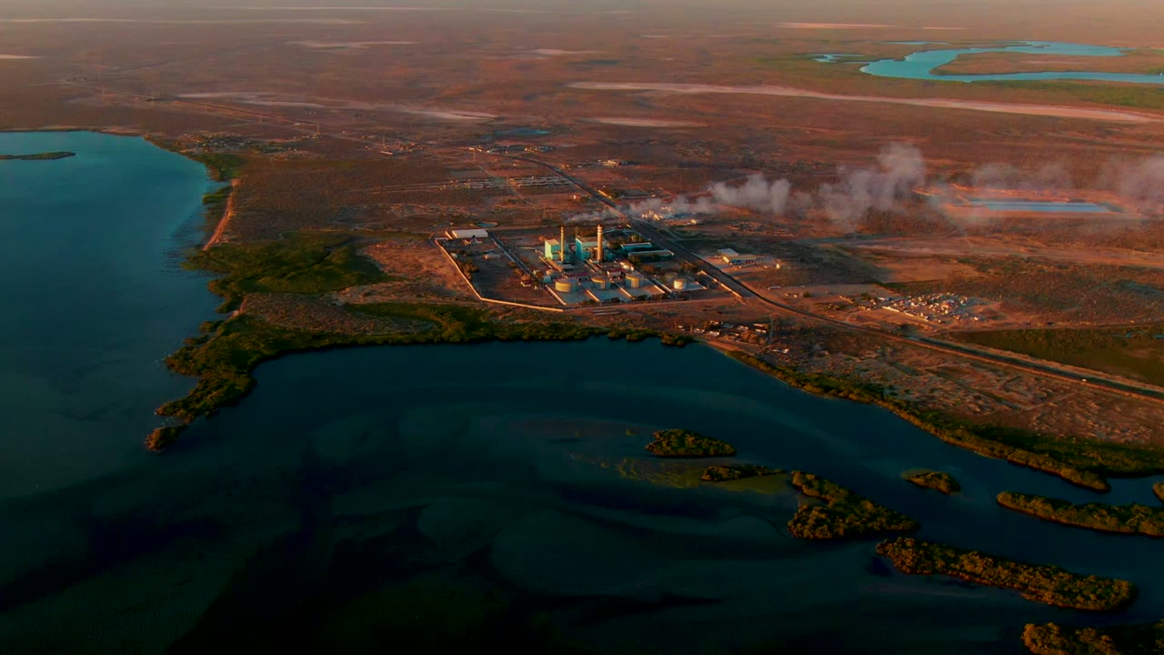 San Carlos, Baja California Sur aerial frame showing the power plant slightly right of center, smoke drifting over brown terrain, turquoise ponds in the distance, and no visible people.