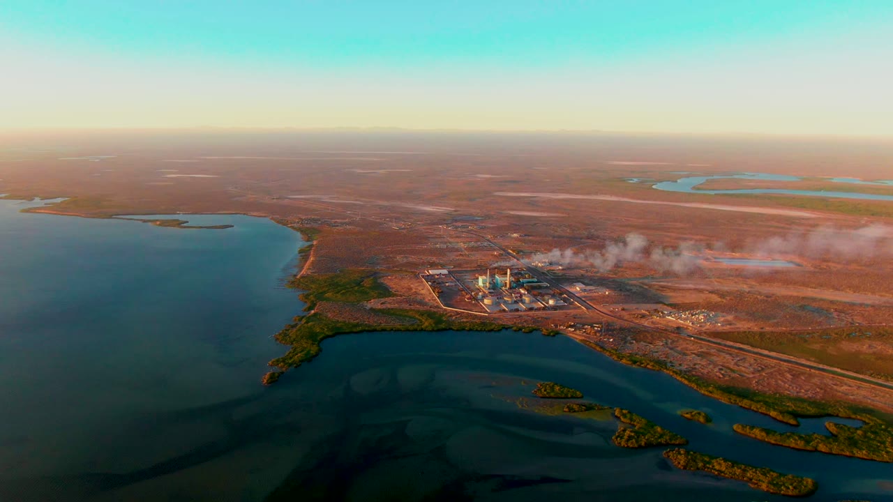 Opening aerial frame over San Carlos, Baja California Sur, showing a blue waterway, brown arid land, an industrial site emitting pale smoke or steam, and no visible people.