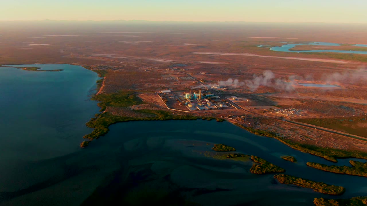 Opening aerial frame of a San Carlos, Baja California Sur power plant with pale smoke drifting over dry brown land beside blue-green water, with no visible people.