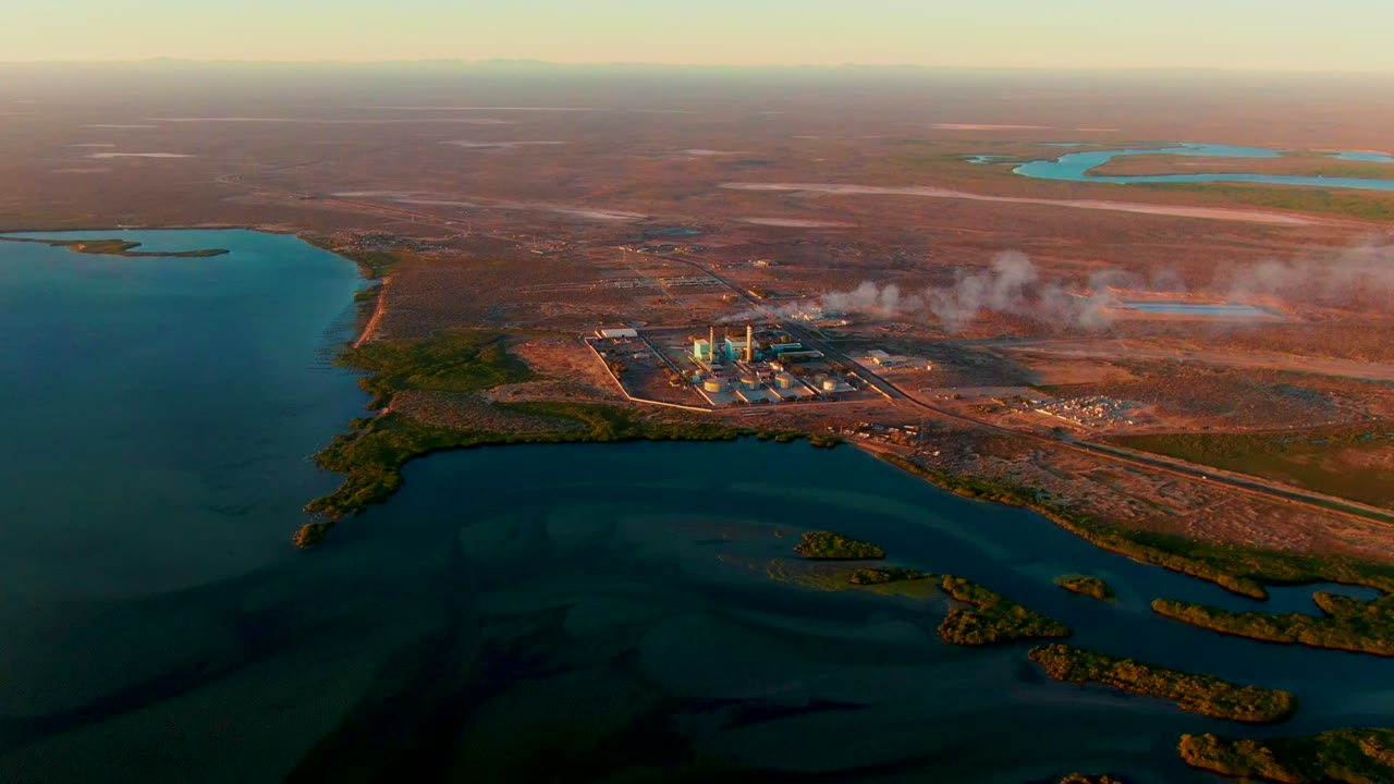 Second aerial frame over San Carlos, Baja California Sur, showing smokestacks and drifting smoke at a power plant between open water and dry inland terrain, with no visible people.