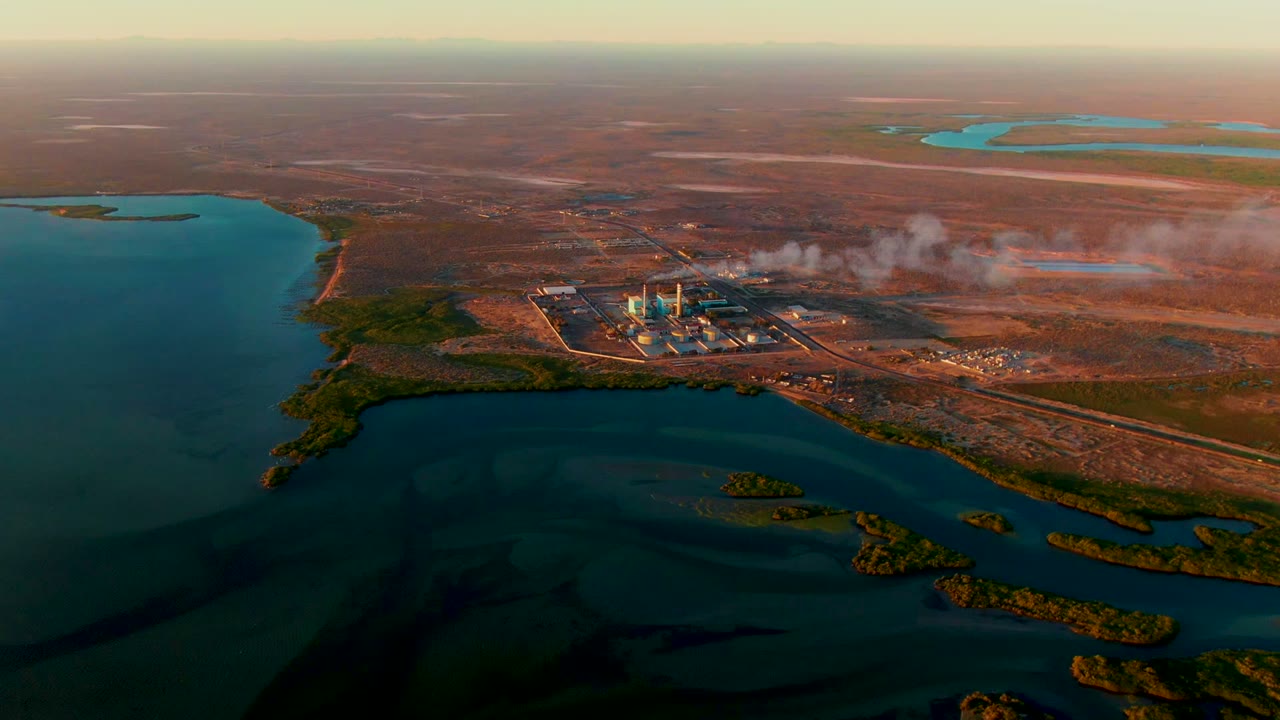 Aerial frame in San Carlos, Baja California Sur with a power plant and white-gray smoke plume on reddish-brown land beside dark blue-green water, with no visible people.