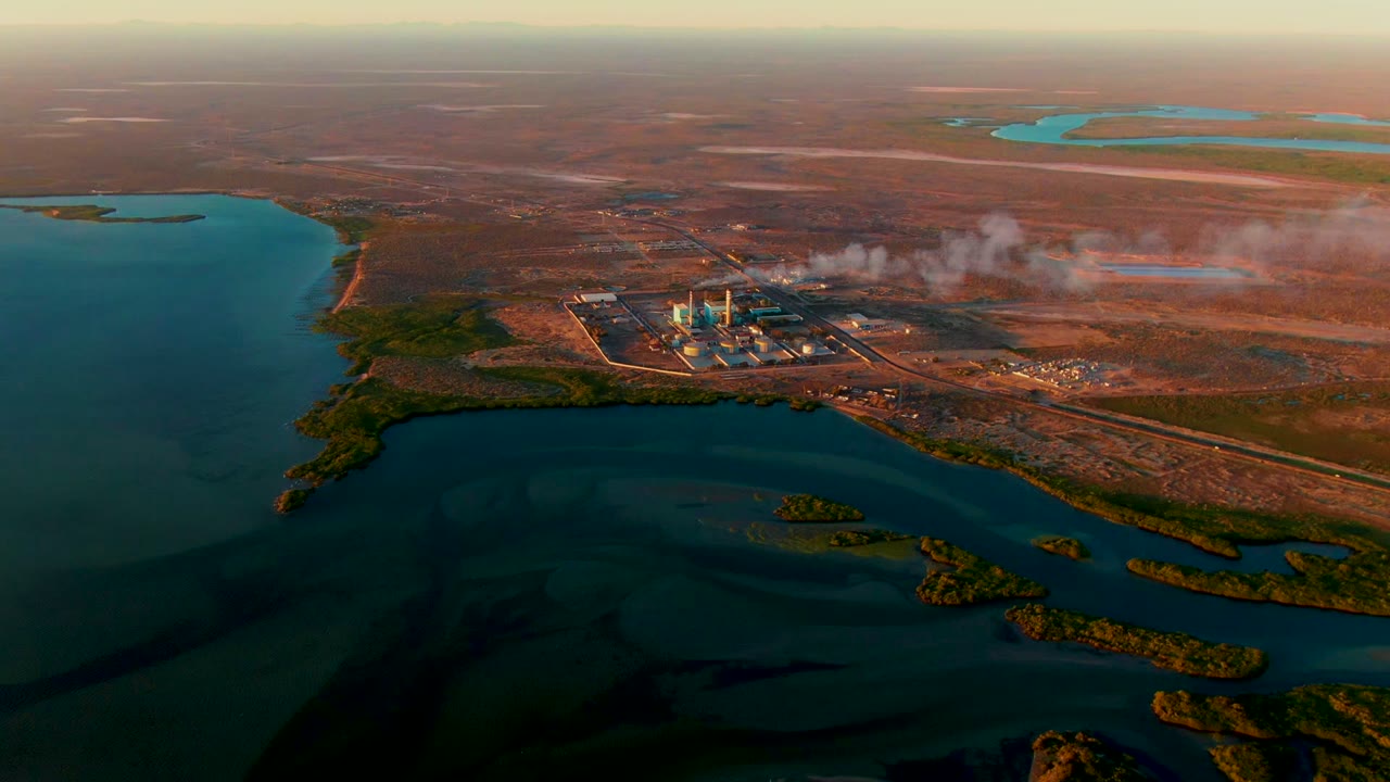 Aerial power plant scene with smoke drifting over dry land and water in front.