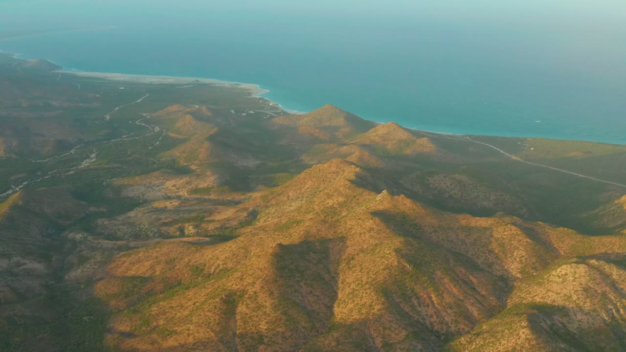 Opening frame over Cabo Pulmo in Baja California Sur, with sunlit ridges in the foreground, a pale shoreline beyond, turquoise ocean at the top, and no visible people.
