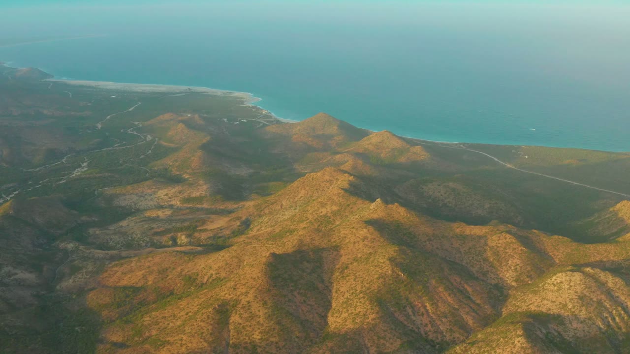 Cabo Pulmo aerial view with a prominent sunlit ridge in the lower frame, darker shadowed terrain to the left, the shoreline crossing the distance, and no visible people.