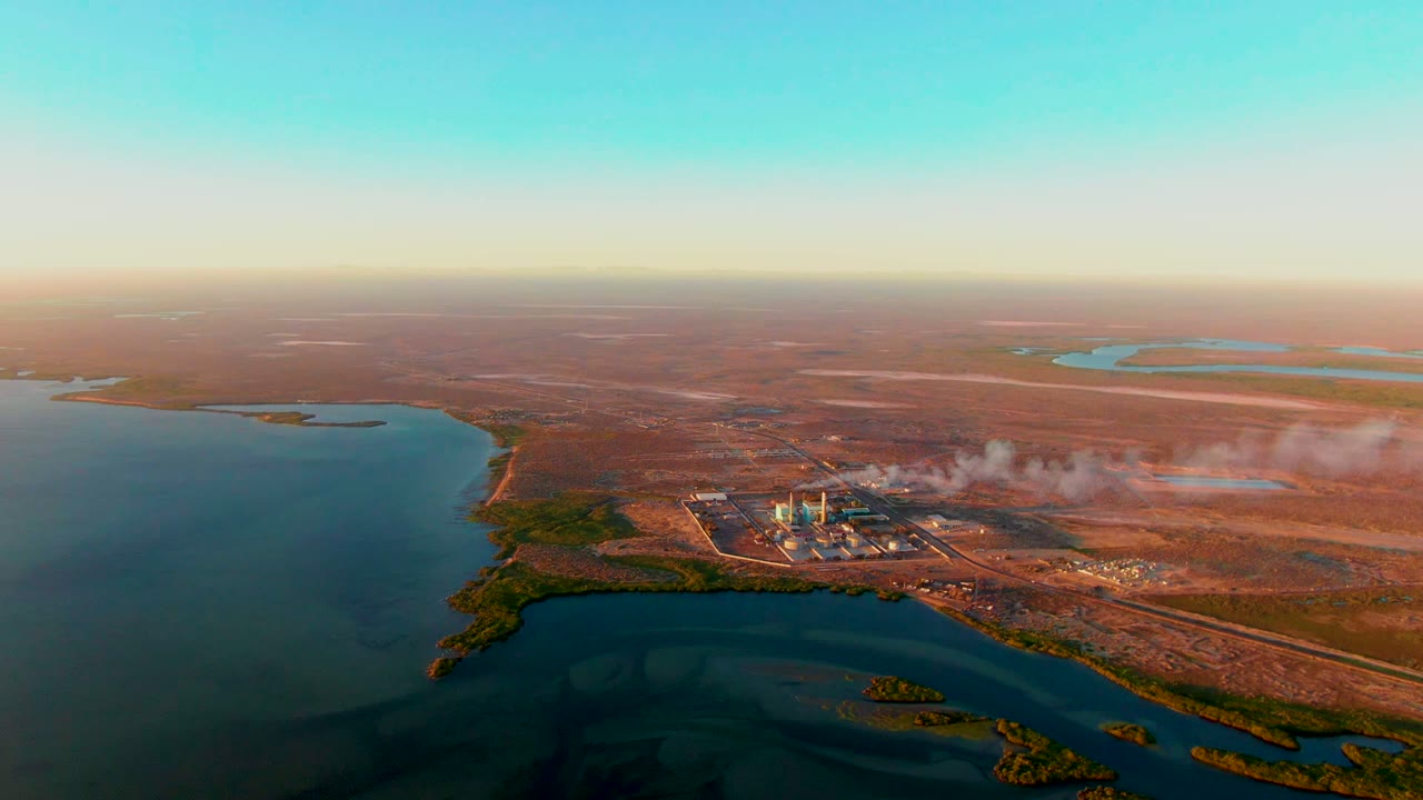 Industrial plant with smoke near shoreline, dry terrain, and distant water channels.