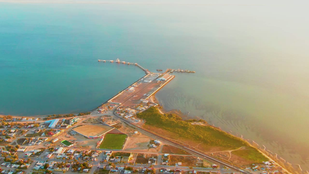 High aerial frame over San Carlos, Baja California Sur, showing a pier projecting into blue-green water, town blocks below, shoreline roads, and no visible people.