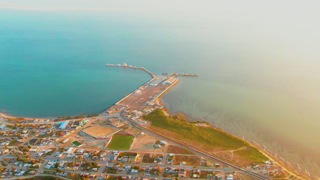 Drone view of a pier, shoreline, roads, buildings, and a green field beside the water.