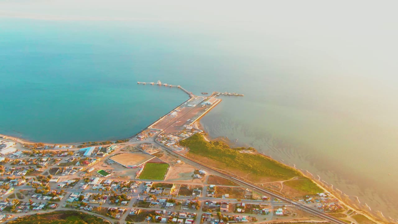 Wide drone frame of San Carlos, Baja California Sur, with turquoise water above the pier, pale shallow water near shore, town blocks at the bottom, and no visible people.