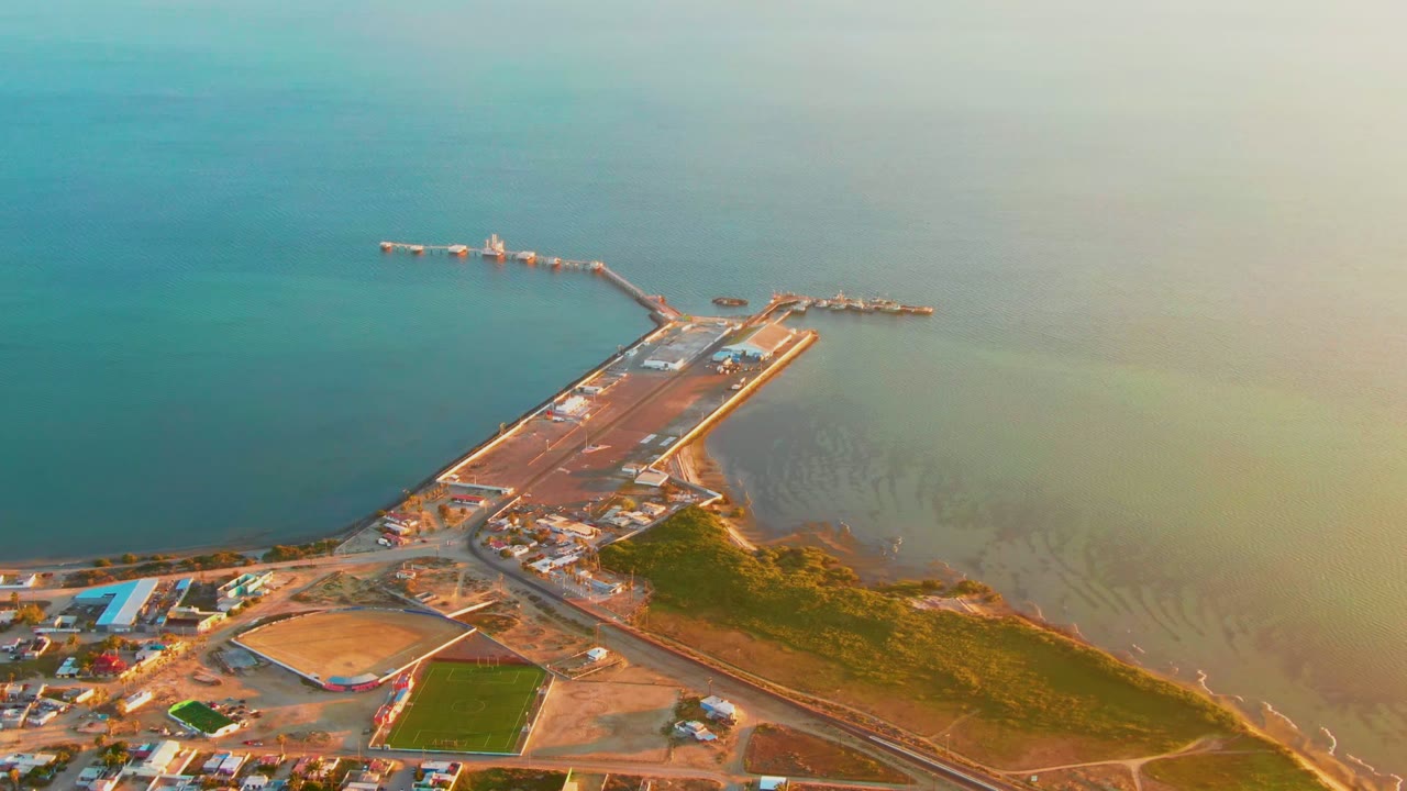 Opening aerial frame over San Carlos, Baja California Sur showing a long pier, small harbor edge, shoreline roads, fields, and calm blue-green water with no visible people.