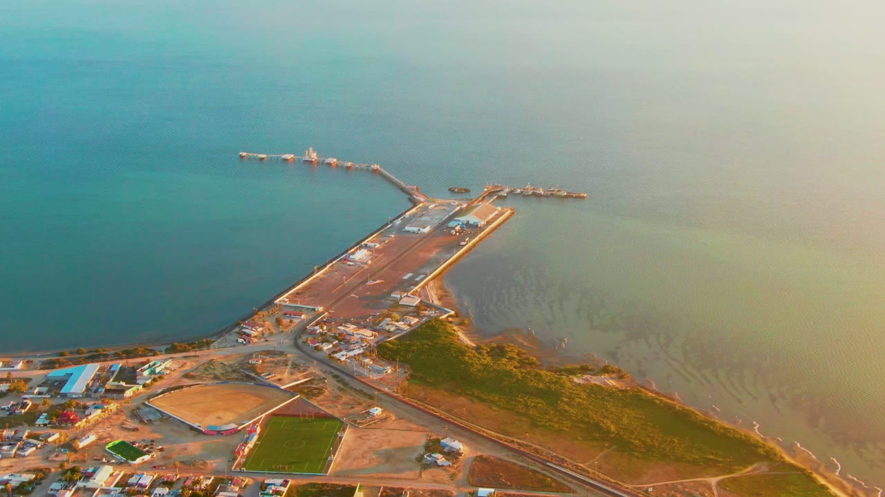San Carlos aerial frame showing the long jetty-like pier, dock area, sandy shoreline development, a green field, and pale water gradients with no visible people.