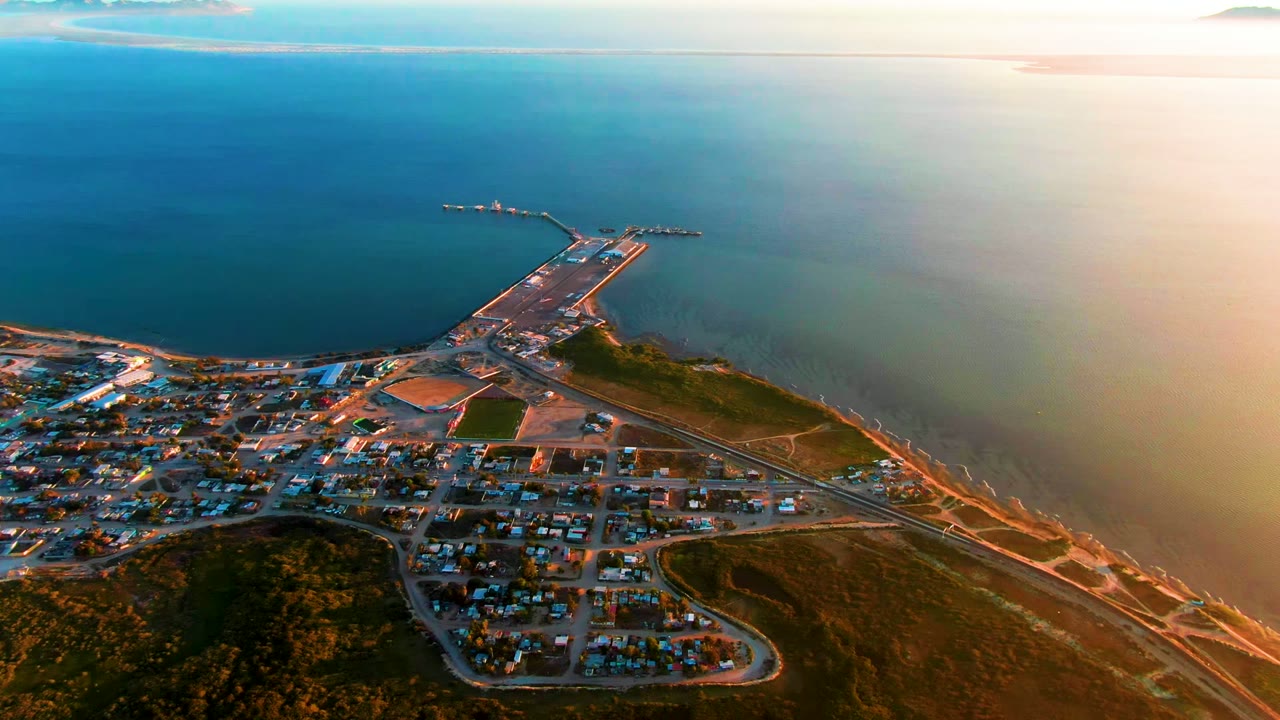High aerial view over San Carlos, Baja California Sur, showing a coastal town below, a long pier extending into blue-green ocean, warm sunset light, and no visible people.