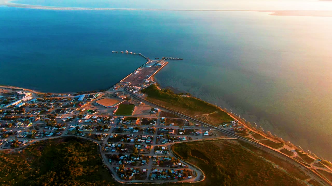 Wide drone view of town roads and a pier beside calm ocean water.