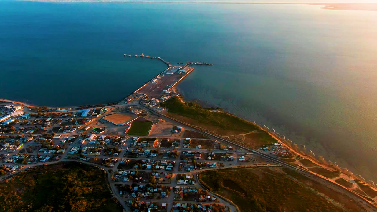 Aerial view over San Carlos, Baja California Sur, with the pier and shoreline slightly higher in frame, ocean water filling the background, orange evening light at the right edge, and no visible people.
