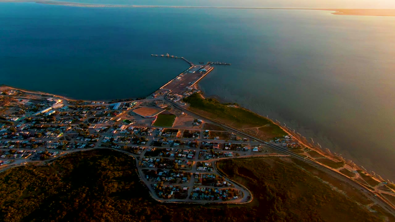 Wide drone view over San Carlos, Baja California Sur, with dark hillside foreground, town blocks, a harbor pier reaching into the ocean, warm sunset light, and no visible people.