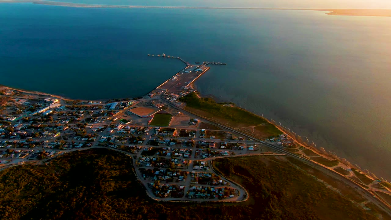 High aerial view of town streets, shoreline work area, harbor pier, and broad ocean.