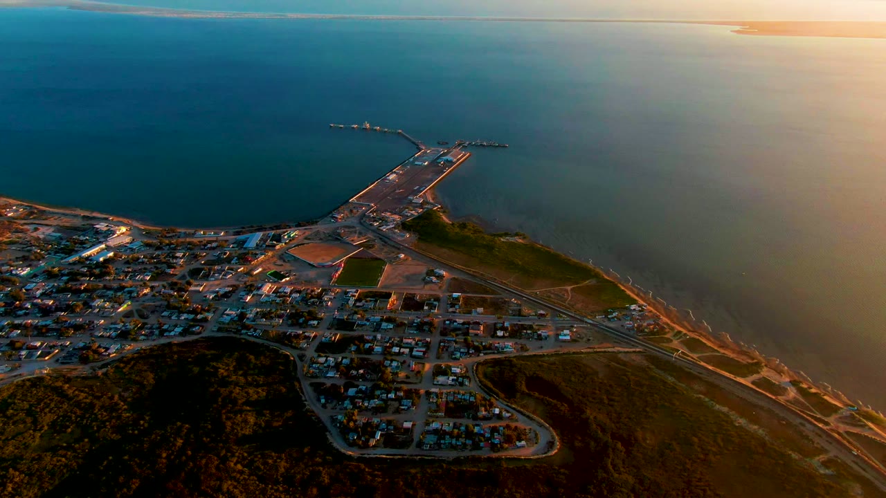 San Carlos aerial view with the harbor pier near the center, town blocks along the lower edge, open ocean across the upper frame, orange evening light, and no visible people.