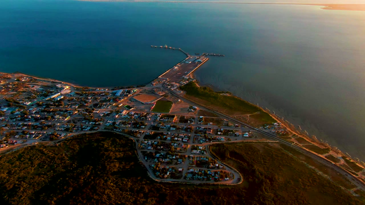A high aerial view over San Carlos, Baja California Sur shows hillside roads, residential blocks, a harbor pier extending into the ocean, warm sunset light, and no visible people.