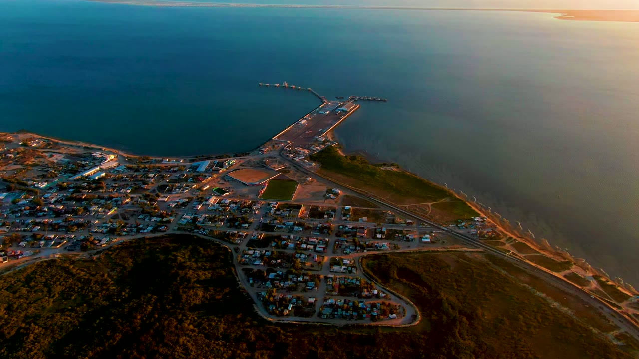 At 0:02, the aerial view over San Carlos shows more open ocean above the harbor, with the pier, jetty, coastal roads, hillside, residential buildings, and no visible people.
