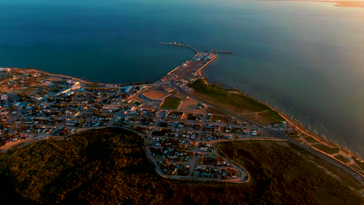 Aerial frame over San Carlos, Baja California Sur showing a dark hillside, town streets, shoreline road, pier extending into calm ocean, and no visible people.