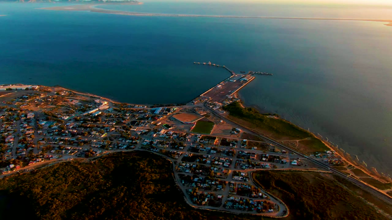 High aerial view over San Carlos, Baja California Sur, with brown hills in the foreground, a coastal town along the shore, a long pier reaching into blue water, and no visible people.