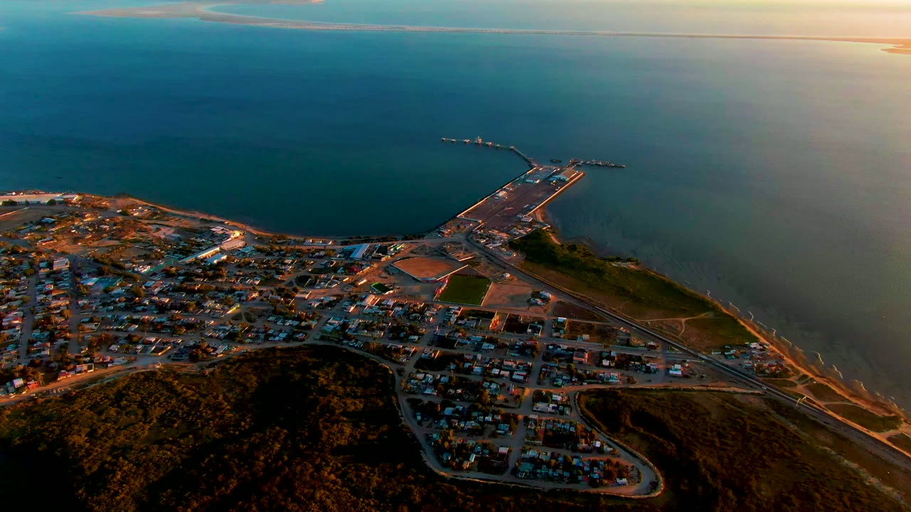 Drone view of shoreline buildings, roads, hills, pier, and calm water.