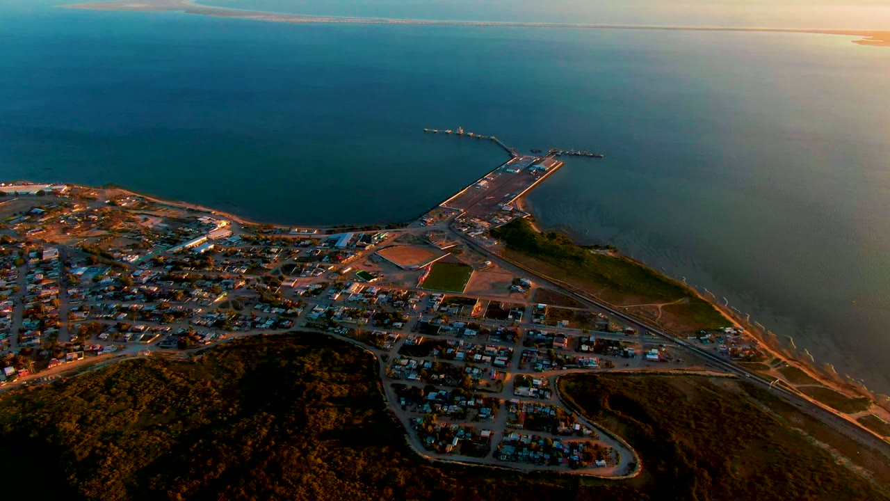 Wide drone view of San Carlos, Baja California Sur, with the town spread across the lower left, hills along the bottom edge, a pier projecting into the ocean, and no visible people.