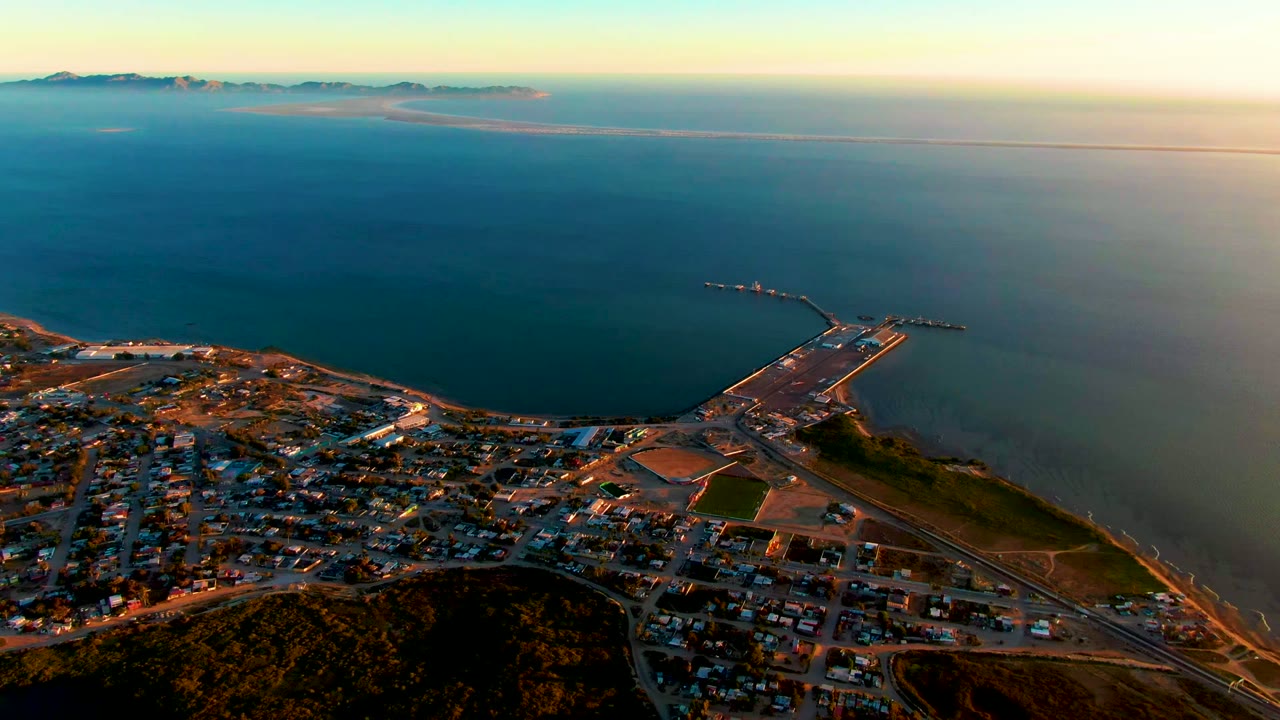 Aerial view over San Carlos, Baja California Sur, with a hillside foreground, town streets below, a harbor pier reaching into blue ocean, distant mountains on the horizon, and no visible people.