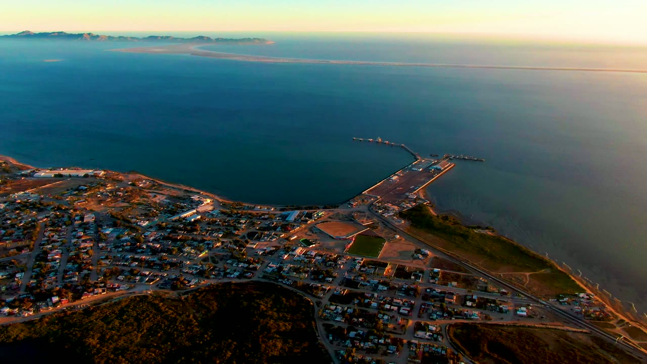 High aerial view of harbor pier and coastal buildings beside a wide blue ocean.