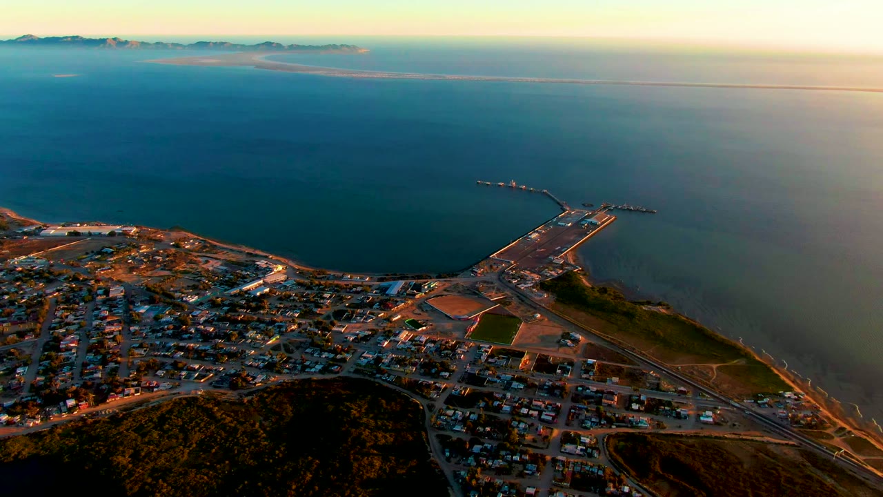 Aerial frame of San Carlos showing more open water around the harbor pier, the town and roads along the lower edge, distant mountains across the ocean, and no visible people.