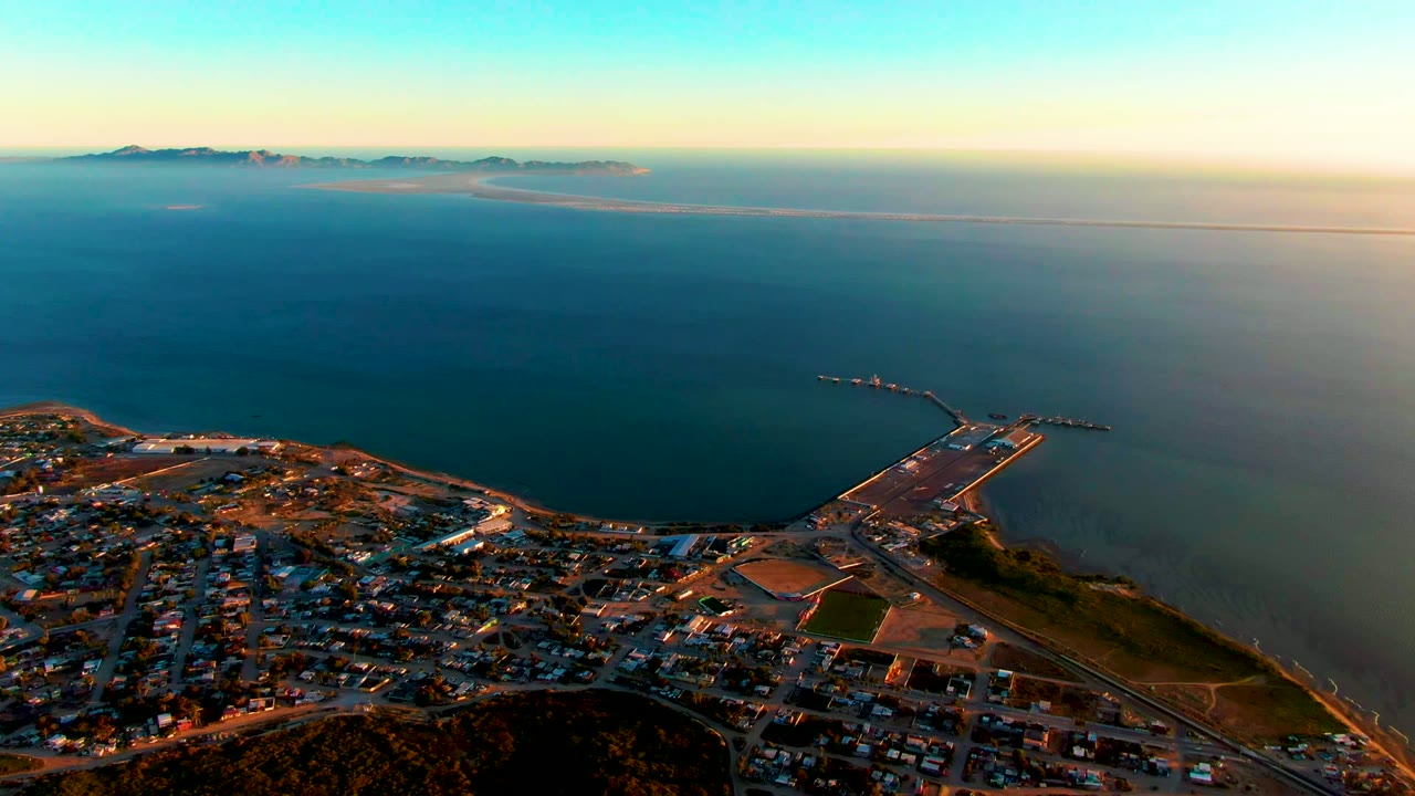 High aerial frame over San Carlos, Baja California Sur showing town streets, low hills, a harbor pier, broad ocean, distant islands, warm sunset light, and no visible people.