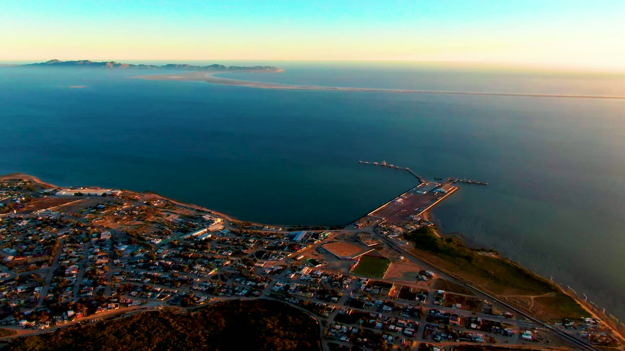 Coastal town and harbor beside calm ocean under golden sunset light.