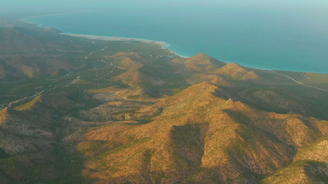 Aerial view over Cabo Pulmo coastal mountain ridges with a blue ocean horizon, warm light on the slopes, and no visible people.