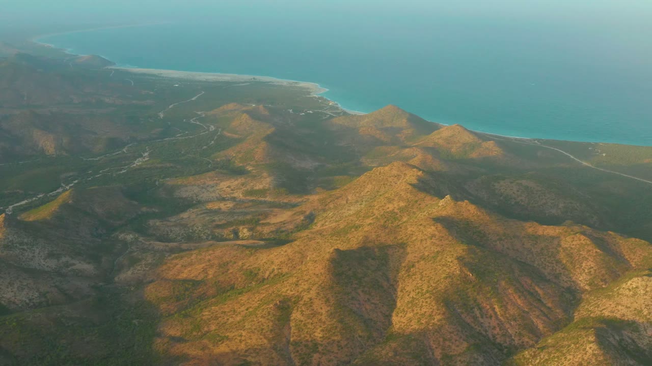 High aerial frame over Cabo Pulmo hills, with a pale road cutting across the terrain and the coastline stretching along the upper background, no visible people.
