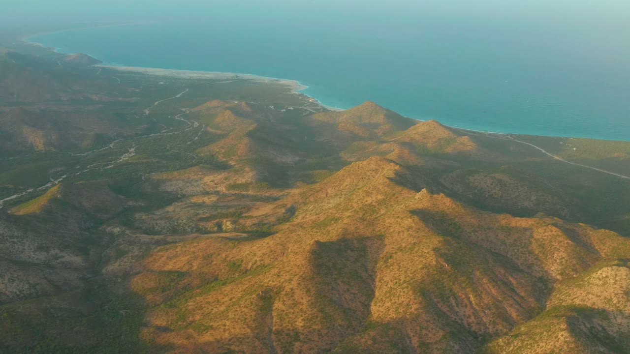 Drone view above Cabo Pulmo with sunlit brown hills in the foreground, darker valleys below, a road line at midframe, and no visible people.