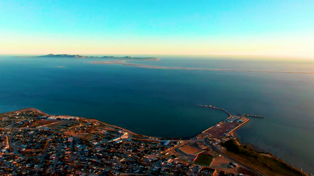 Aerial view over San Carlos, Baja California Sur with town buildings at the bottom, calm ocean across the frame, a pier at right, distant landforms on the horizon, and no visible people.