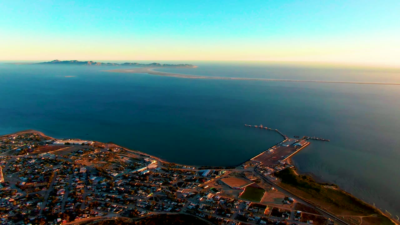 Elevated view of San Carlos, Baja California Sur with dense town blocks in the lower left, a pier and paved waterfront area at right, open ocean in the middle, distant landforms, and no visible people.