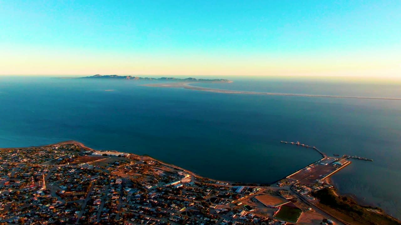 Aerial view over San Carlos, Baja California Sur, with town streets along the lower frame, blue ocean beyond, a breakwater at right, distant land on the horizon, and no visible people.