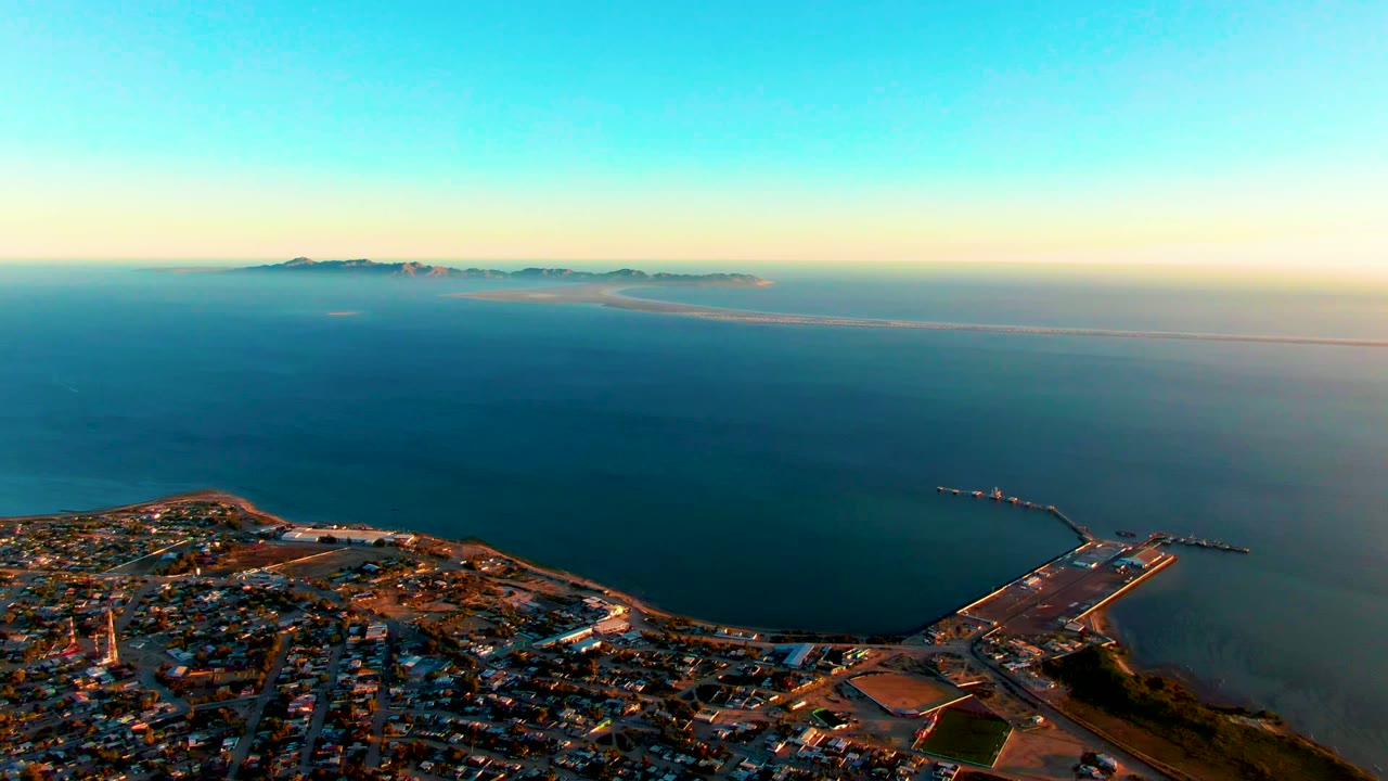 Aerial view of San Carlos, Baja California Sur, with the town and road grid at the bottom edge, a broad expanse of blue sea, a harbor structure at right, distant land, and no visible people.