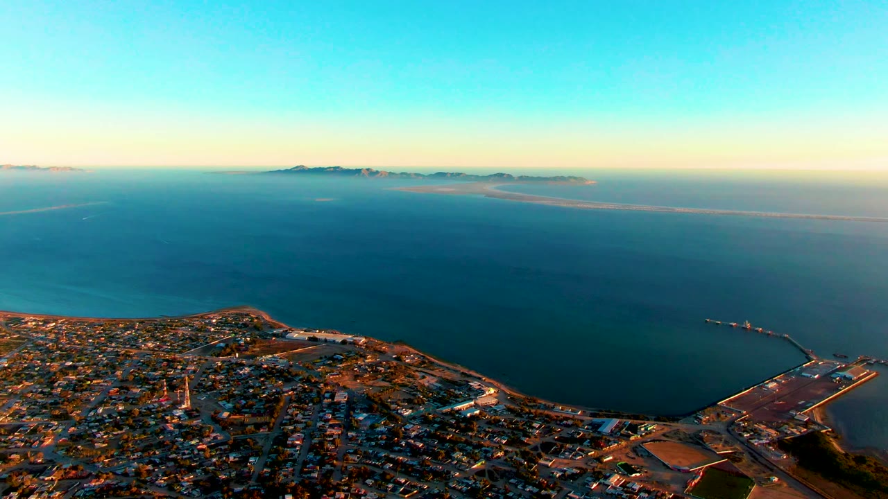 Opening aerial frame over San Carlos, Baja California Sur, with city blocks along the lower edge, a port at lower right, broad blue ocean, distant islands on the horizon, and no visible people.