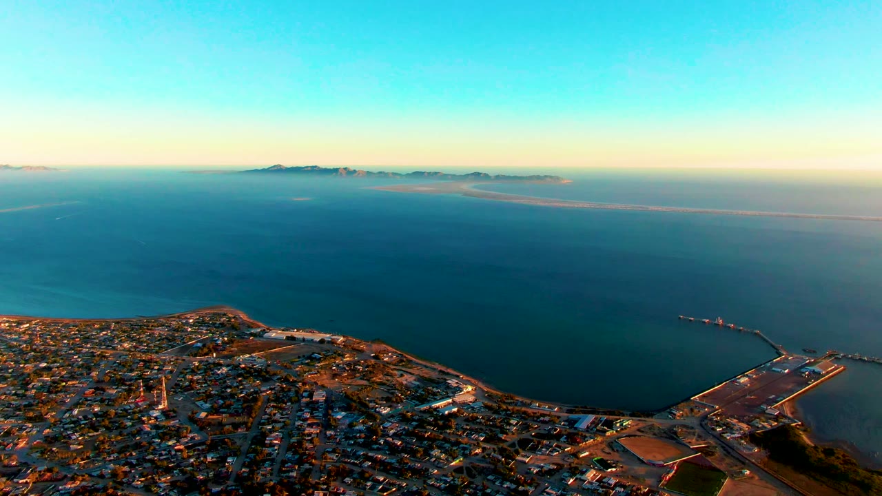 Wide San Carlos aerial with dense low buildings at the bottom, dock and port geometry at lower right, smooth blue water in the middle distance, distant islands, and no visible people.