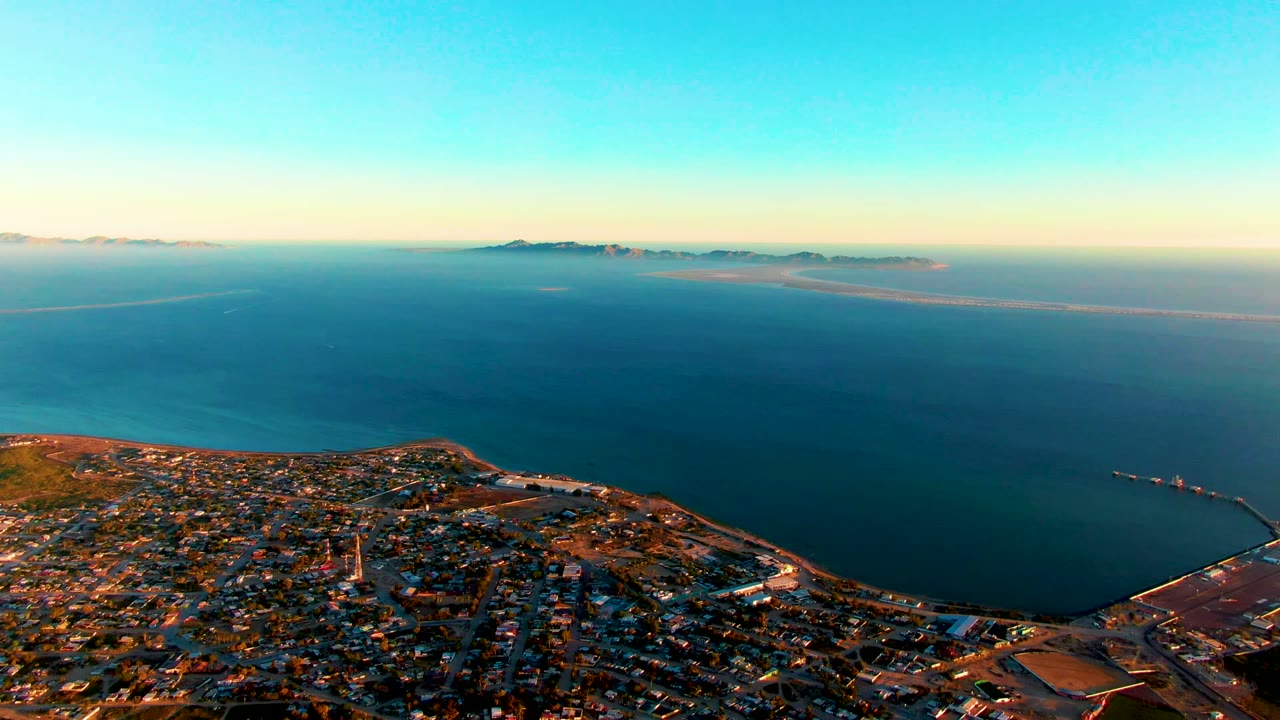 Opening aerial frame over San Carlos, Baja California Sur, with dense coastal buildings below, calm ocean beyond, distant islands on the horizon, and no visible people.
