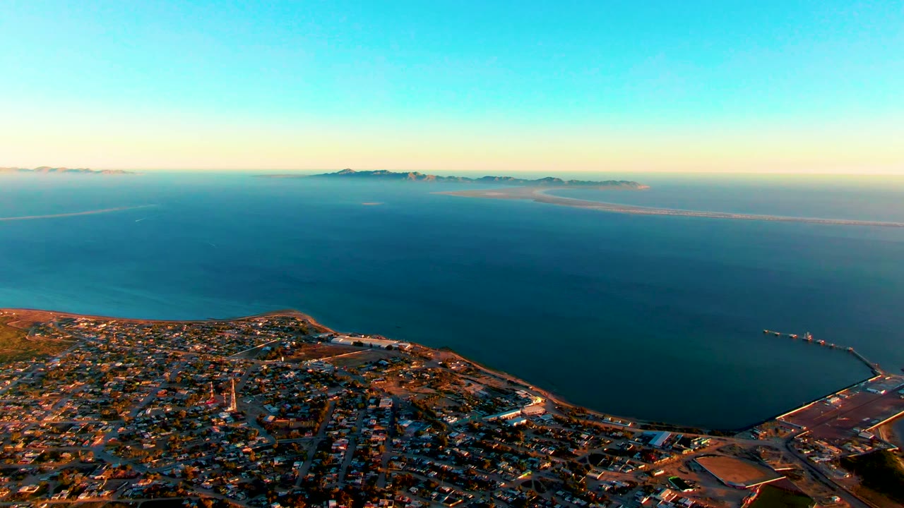 Third frame shows San Carlos from above with a harbor edge and breakwater at lower right, dense buildings along the coast, smooth ocean water, distant islands, and no visible people.