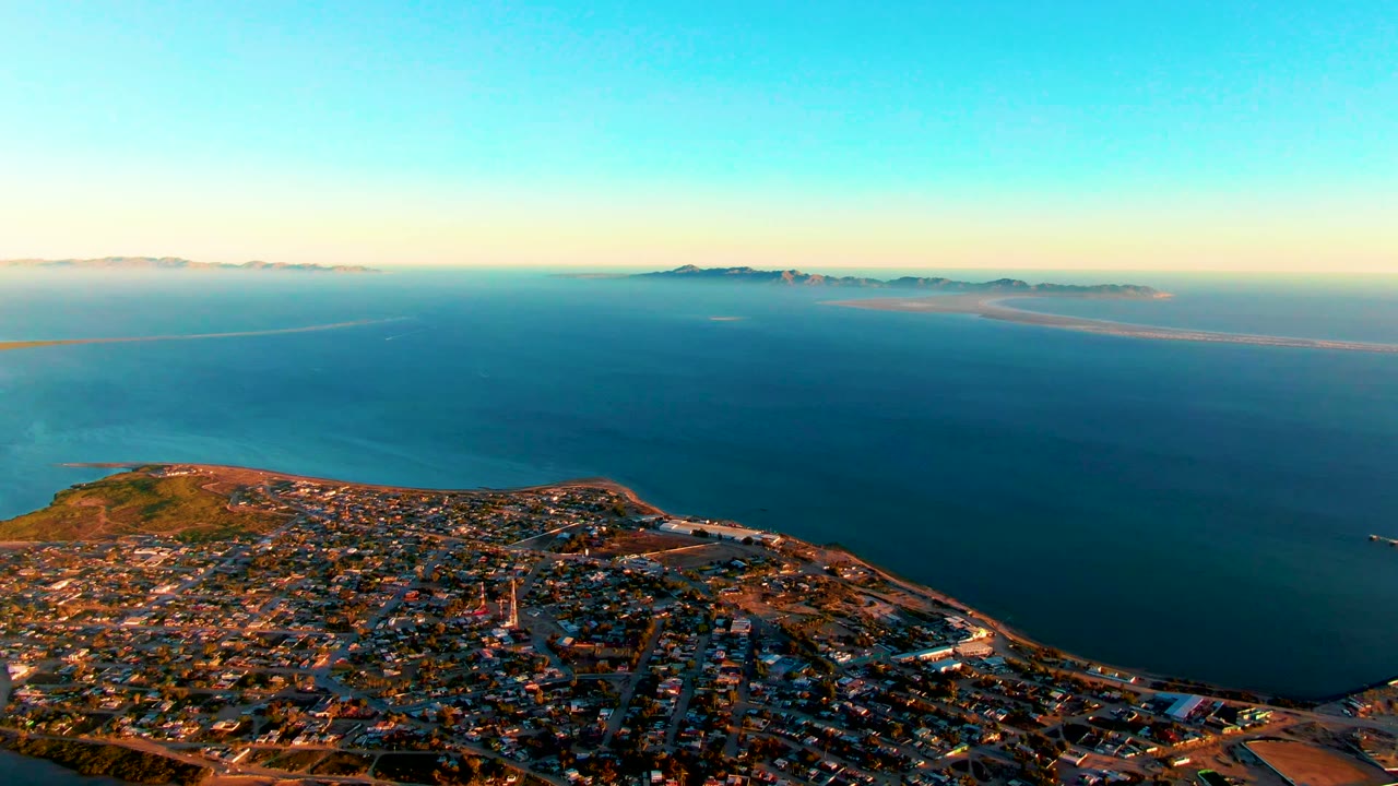 Wide aerial frame over San Carlos, Baja California Sur, showing dense buildings at the bottom, calm ocean across the center, distant islands on the horizon, and no visible people.