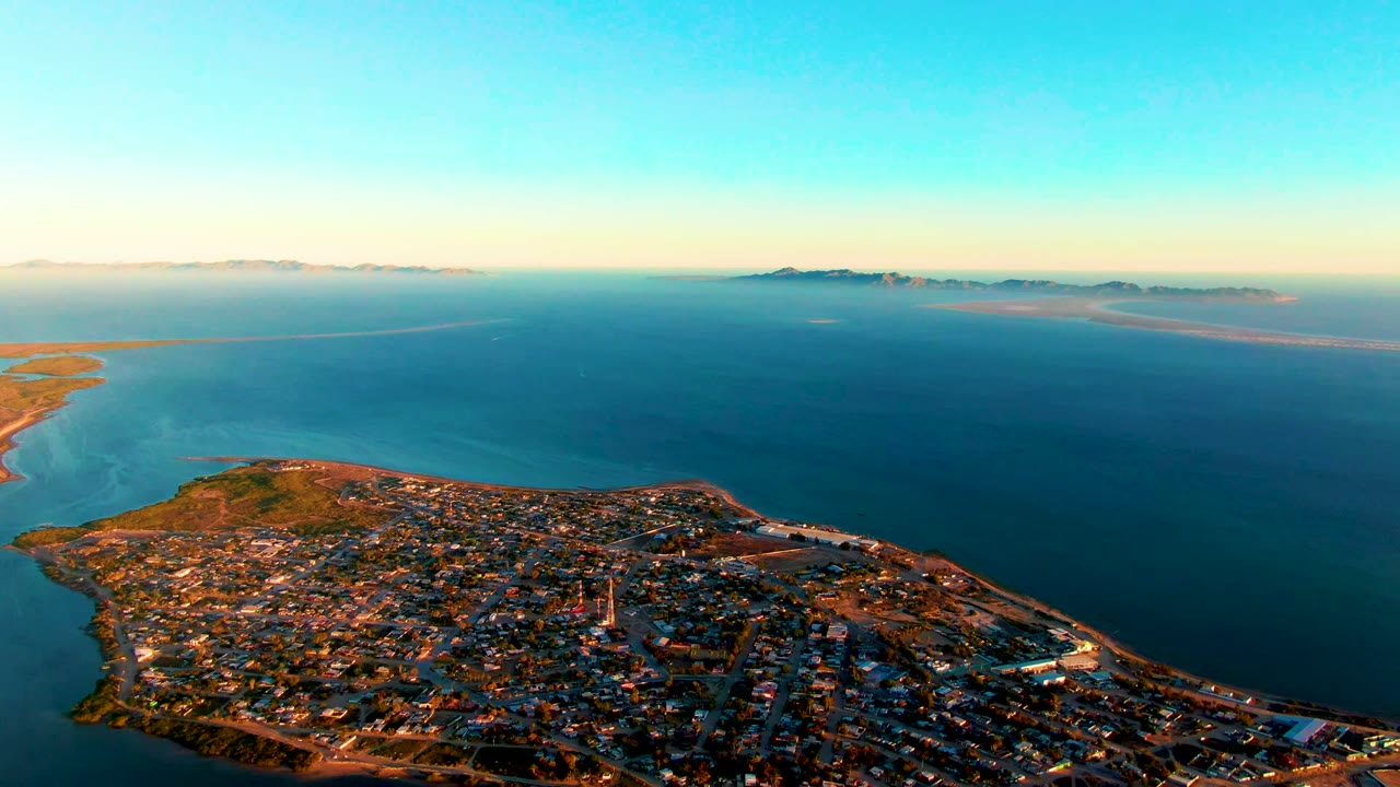 Opening aerial frame of San Carlos, Baja California Sur, with the town spread across a peninsula, calm blue water around it, distant islands on the horizon, and no visible people.