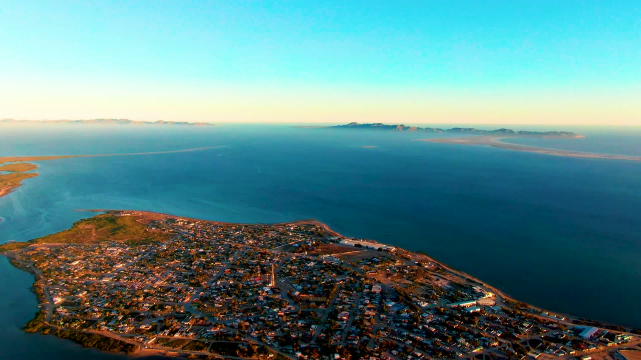 Wide drone view over rooftops and shoreline with open sea beyond.