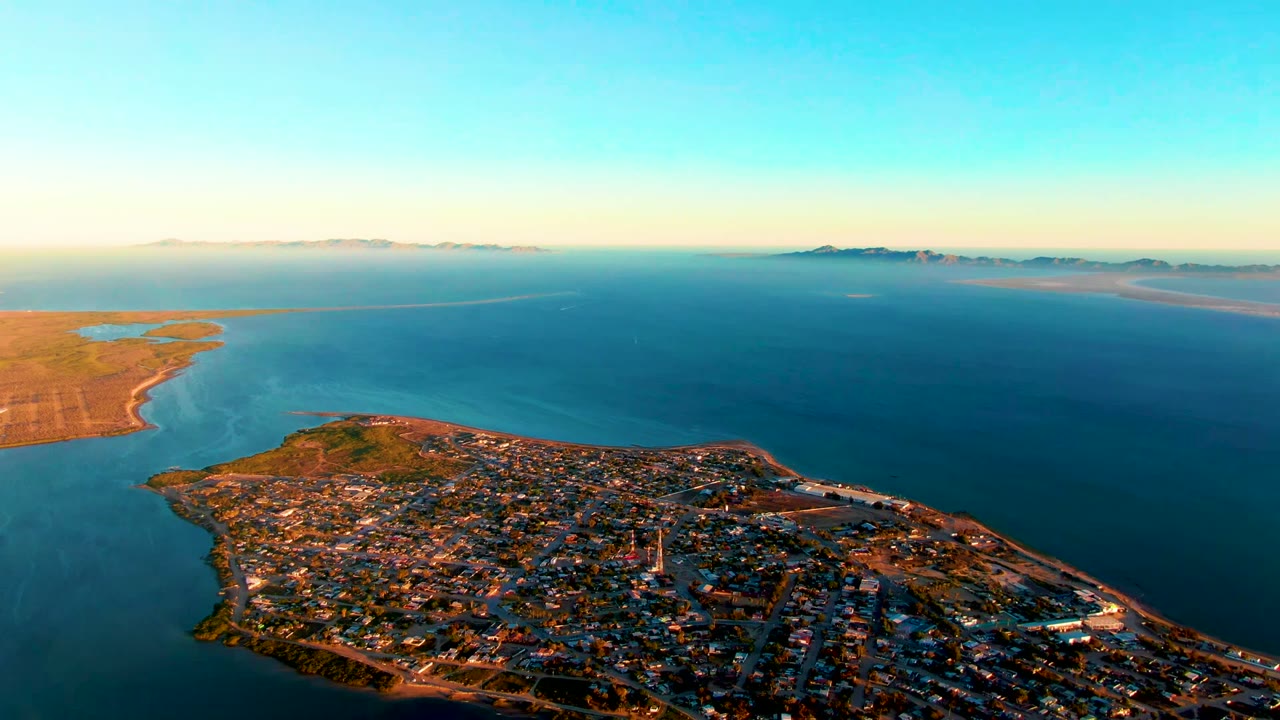 High aerial frame over San Carlos, Baja California Sur, showing the town filling the lower peninsula, blue sea around it, distant land on the horizon, warm sunrise light, and no visible people.