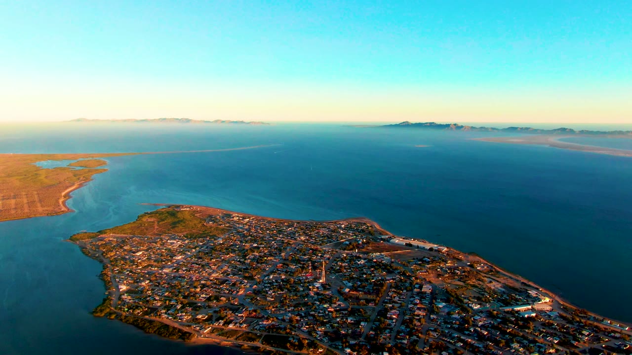 Aerial view of San Carlos with the town grid across the lower foreground, a curved shoreline at left, broad blue water through the center, distant landforms, and no visible people.